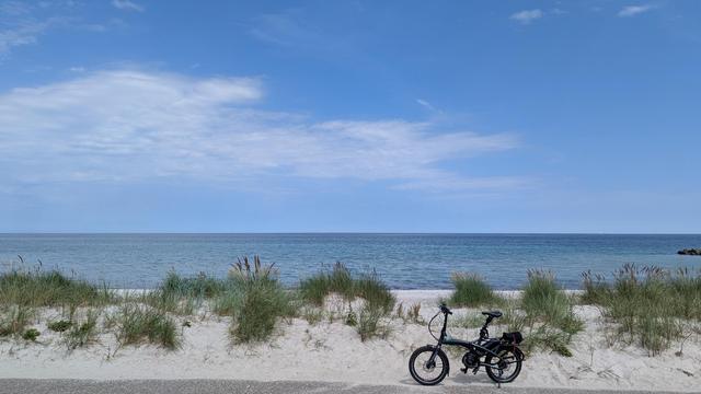 Blick auf die Ostsee vom Deichfuß aus. 
Dünen mit Strandhafer-Bewuchs begrenzen den Deich vom dahinterliegenden Strand. Vor den Dünen steht ein Tern Faltbike. 
Im Hintergrund ist die blaue Ostsee und der blaue Himmel mit einigen dünnen Wolken.