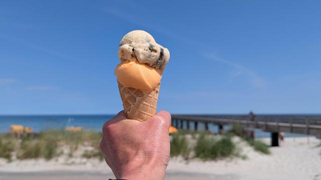 Eine Hand hält ein Eis in einer Waffel hoch. 
Dahinter ist verschwommen der Strand, eine Seebrücke und das Meer.