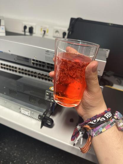Ein glass mit alkohol freiem Aperol Spritz, gehalten von einer Hand mit diversen Festivalbändern vor einem Stapel Ethernet Switche