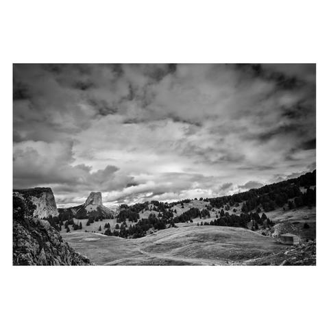 Photo en noir et blanc d'un paysage de moyenne montagne.
La partie basse de l'image est occupée par un plateau herbeux vallonné, un peu plus loin il est parsemé d'épicéas. Sur la gauche on distingue des falaises de faible hauteur, et au fond un sommet, le Mont Aiguille, évoquant le relief de Monument Valley. Dans le coin inférieur droit se dresse une curieuse petite cabane de forme octogonale.
Les deux tiers supérieurs de l'image sont occupé par un ciel dramatique.
-
A black-and-white photo of a mid-mountain landscape.
The lower part of the image is taken up by a rolling grassy plateau, dotted with spruce trees a little further out. On the left, low cliffs are visible, and in the background, a peak—Mont Aiguille—that evokes the landscape of Monument Valley. In the lower right corner stands a curious little octagonal cabin.
The upper two-thirds of the image are filled with a dramatic sky.