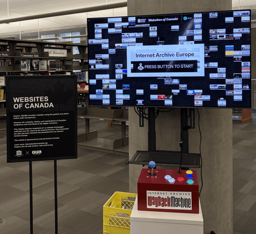 The "Websites of Canada" interactive kiosk installed at the Vancouver Public Library, consisting of a large display screen showing a mosaic of archived Canadian website screenshots and an Internet Archive Wayback Machine arcade-style controller box with colorful buttons and a joystick, mounted on a yellow milk crate. An explanatory signboard stands to the left. The screen prompts visitors to press a button to start, presented by Internet Archive Europe.