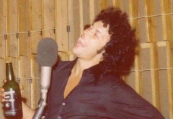 Grainy 1974 photograph of Tim Curry, a white man with dark curly hair. He's wearing a black shirt and is in a recording studio. There's a large microphone in front of him. There's a bottle of port in his right hand, and Tim is mid song, head thrown to the side and mouth wide open as he belts something out in that way he has.