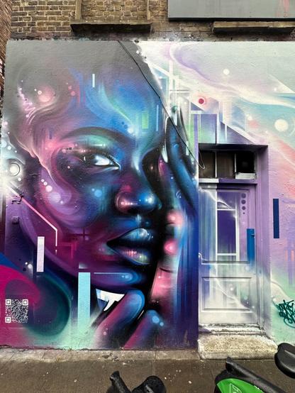 a wall and door at street level, painted with an intricate, colourful mural with a futuristic/scifi aesthetic, featuring a dark-skinned, hairless person’s face illuminated with blue and purple light, alongside pastel-coloured geometric elements