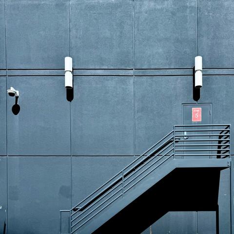 Back of an all black building with a black exterior stairwell leading up to an all black door with a red son on it, which is kind of the only indication that the photo is not in black and white.