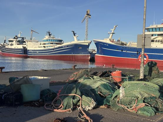A harbor scene featuring several large fishing boats docked at a wharf. In the foreground, there are piles of fishing nets and equipment. The sky is clear, and the water is calm.