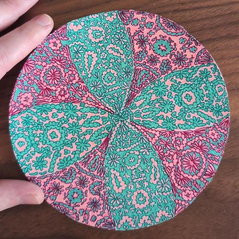 Circle-shaped paper cut-out with intricate patterns, mainly petals and swirls,  drawn on it using fineliner pens in the colours burgundy red, coral pink, teal and dark green. Background is a walnut wood table. You can also see my index finger and thumb in the left corners. The cut-out is roughly the size of my hand.