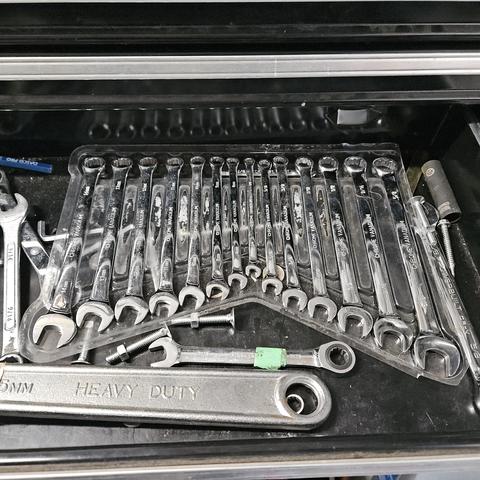 a black tool box drawer is open and exposing a full set of open end, closed end wrenches completing a preformed clear plastic packaging. They are chrome and reflecting warbled environment lighting.