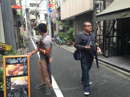 Om Malik walking the streets of Tokyo, wearing all black clothes, sneakers and carrying a shoulder bag, umbrella, and a Leica.