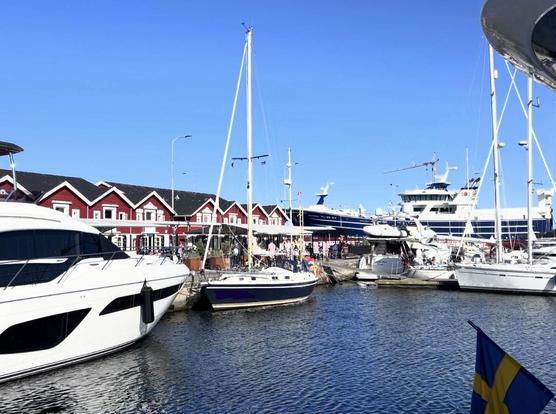 A picturesque harbor scene featuring several boats and yachts docked along a waterfront, with charming red buildings in the background and a clear blue sky overhead.