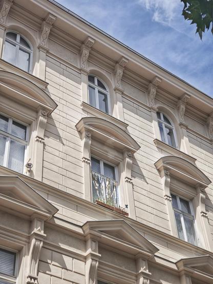 Das Foto zeigt die Fassade eines mehrstöckigen, hellen Altbaus aus einer schrägen Perspektive von unten nach oben. Das Gebäude hat viele architektonische Details: verzierte Gesimse, dekorative Konsolen, Pilaster und mehrere Fenster mit geschwungenen Überdachungen. Die Fassade wirkt klassisch und gepflegt.

Die Fenster sind geschlossen und haben Jalousien auf der Innenseite. Auf einer Fensterbank steht ein kleiner Blumenkasten. Vor diesem Fenster flattert eine Reihe gold-silberner Folienstreifen. Oben rechts sieht man einen Ausschnitt von Baumblättern vor einem blauen Himmel mit leichten Wolken.