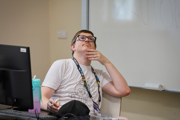 A person sits at a desk in front of a computer monitor and a large whiteboard. The individual wears black-rimmed glasses and a white t-shirt featuring a line drawing of a character's face. A lanyard with blue, yellow, and black patterns hangs around their neck. They have one hand resting against their chin while looking upward toward the top right corner of the frame. To the left, there is a black monitor next to a pink container holding pens.
