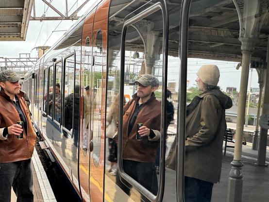 a London Overground train standing at a platform, both its windows and metal surfaces polished to perfection and reflecting the abundant daylight. Passengers stand, awaiting the opportunity to board. The reflection of one passenger, a man in a cap holding a can of Guinness, is framed in a train window