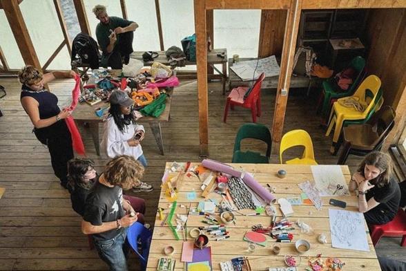 Vogelperspektive auf einen offenen Workshop-Raum mit Holzdielen: Mehrere Personen arbeiten an einem langen Holztisch voller Bastelmaterialien wie Stoffbänder, Stifte, Papier und Werkzeuge. Eine Person hält ein rotes Stoffband hoch, andere sitzen oder stehen im Gespräch. Im Hintergrund stapeln sich bunte Plastikstühle, ein weiterer Arbeitstisch mit Material steht links im Bild.