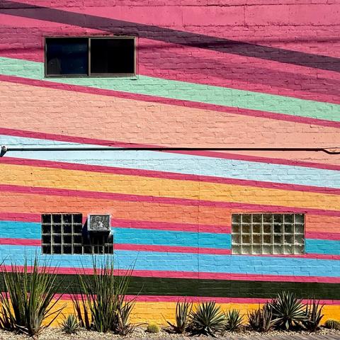 Side of a building painted in a decorative mural featuring diagonal lines in a variety of colors like yellow, orange, pink, mint green, and blue. On the side are also three windows. One upper very normal window and two lower glass block windows where one was an air conditioner in it.