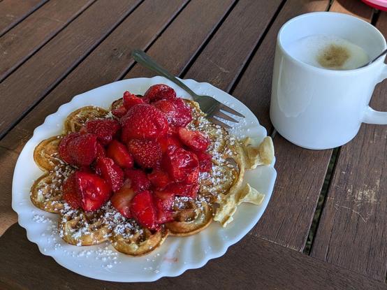Auf einem Holztischn ein Teller mit Herzchenwaffel und einem Haufen Erdbeeren darauf, dahinter eine Tasse Cappuchino.