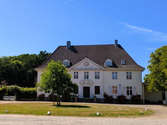 Das Gutshaus Steinwehr, ein mehrstöckiges Gebäude mit großer Auffahrt um ein rundes Rasenstück davor. Dahinter Bäume und blauer Himmel mit einem Schleierwölkchen.