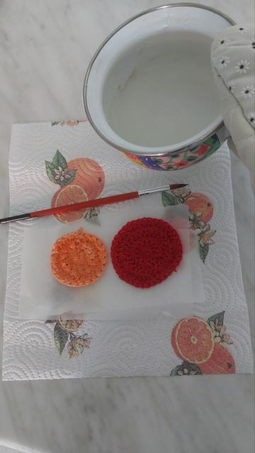 Two crochet circles (one orange, one red) sitting on a sheet of baking paper layered over kitchen paper with a citrus print. A brush rests above the circles. A white pot containing a pale gel is held from the side.