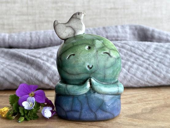a raku ceramic jizo nature spirit guardian sculpture glazed in a bottom-to-top gradient of blue to forest green to spring green. they have a happy, smiley face, little praying hands, and a sweet white bird on their head. they're standing on a piece of chestnut wood with grey muslin and natural linen behind. next to them on the left are a rule rock cress flower, some speedwell (green leaves and tiny white-indigo-violet flowers), and a yellow hawkweed flower.