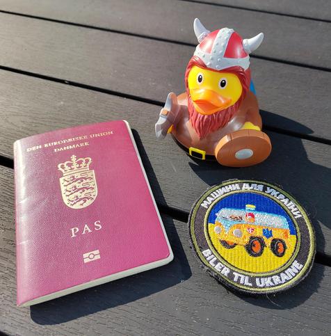 Photo of three items on a table. One beetroot colored Danish passport, one velcro chevron entitled "Biler til Ukraine" (Cars for Ukraine) and one rubber duck with viking helmet, battle axe, shield and a red beard. All items that I bring on the road with me.