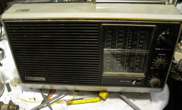 A Pye 1780 portable radio tuned to Radio 4 Long wave, on a  rather messy bench. The radio is from the late 70s, and has a grey case with black speaker grill and frontage, a vertical scale with clear plastic cover over the scale and some knobs with orange highlighting.