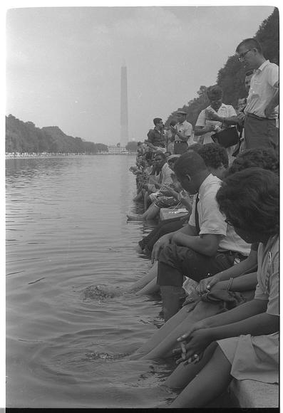 Photo of  dozens of people sitting along the edge of the Reflecting Pool on the Mall in Washington D.C. They are comfortably resting with their legs and feet in the water. The photo was taken during the 1963 March on Washington. It is from the Library of Congress photo collection and is in the public domain.