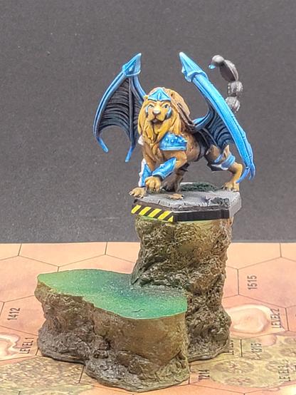 A fantasy miniature of a Manticore, a beast with the body of a lion, the wings of a bat, and the tail of a scorpion, perched atop a battletech hex base, with the front of the base, with its hazard stripes, showing.  The view is a slightly elevated front right view, allowing a somewhat skewed view of the base, which is an urban concrete surface, nice and flat, with a fallen bit of sheet metal fence, largely rusted, and a torn up teddybear beneath the manticore's raised left paw.

The manticore itself is a bright golden brown fur, with a blue metal tiara-like helmet with wings protecting its jaw, metallic vambraces wrapped around its forelegs, one in blue metal, while the other is blue metal with a white stripe, indicating that it is part of the Lyran Guards.  The shoulders similarly have spiked metal pauldrons, blue metallic, with the right having a white stripe on the inside.

The wings are a bright cyan with darker vaguely purple-blue depths, with the upper tips of the wings guarded with that same blue metal, as well as a knee guard on the rear leg, held on with a cloth loop of some sort, and the blue leather belt holding something on its back.

The manticore's lion-like face has its jaw closed, and it looks in a dignified manner at the camera, except for its left eye seeming to have a raised eyebrow, making the eye noticeably larger than the right eye.