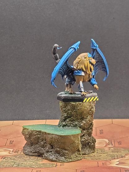 The front left view, showing off the brilliant white stripe of the Lyran Guards on its armor, and some grass having managed to grow through cracks where a large rock has landed during a previous battle or damage.  The brown tail with metallic blue spike is visible from this angle, as well.
