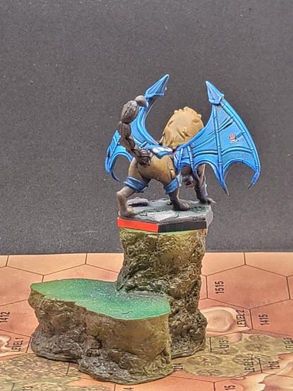 A rear left view, showing off the tail, and the white armor stripe continuing down some padded back armor, being held on by the belly band.  The rear of the wing is a brighter blue, the folds in the wing more obvious, and on its outer section is the Lyran Commonwealth logo, a bronze mailed fist.