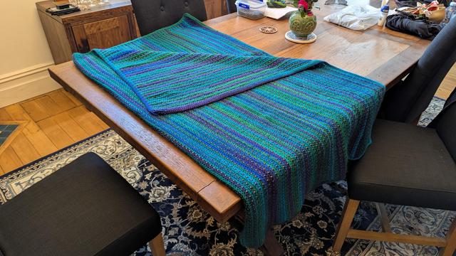 A blue & green multi-colored crocheted blanket spread on a table. Shiny beads a woven into it.