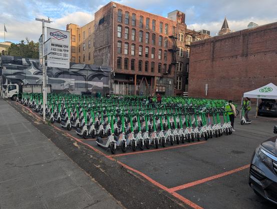 Many, many, MANY green and white Lime scooters and bikes neatly arranged in a parking lot turned over to their use. The buildings are all old brick in the Pioneer Square neighborhood of Seattle, just a few blocks from the stadium where a World Cup game was underway.