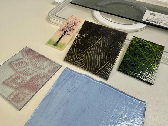 Small glass pieces in various colours as experiments for fusing: one small light pink rectangle with fused chips to resemble a sakura blossom tree, sgraffito paint experiments depicting leaves, a transparent piece with a doily pattern made by powder through a stencil, and another stencilled geometric piece