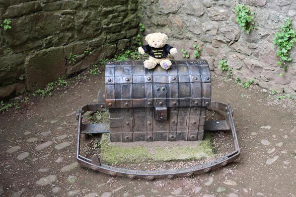 A small blond bear sits on the top of a chest, a trap is set around the chest.
The whole apparatus sits on a block floor, surrounded by stone walls, weeds grow in the gaps in the walls.