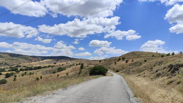 weisse Woljen, blauer Himmel, karge gelb verbrannte Hügellandschafr, vereinzelt grüne Büsche, eine schmale Strasse