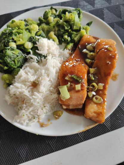 2 pieces of fried salmon, glazed with honey/garlic sauce served on a plate along side white rice, broccoli mixed with spinach and green onions. Green onion also sprinkled on top of the salmon pieces.