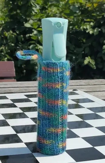 Brumisateur cylindrique dans son étui tricoté arc-en-ciel à dominante bleu-vert sur une table à damier d'un parc public. En arrière-plan, du feuillage.