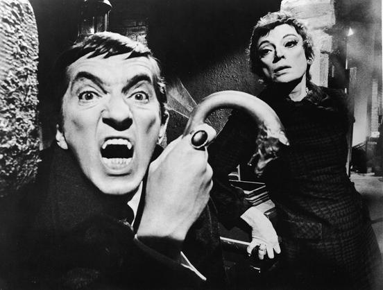 Black-and-white image of Jonathan Frid as Barnabas Collins, dressed in black, holding a cane with a wolf's head handle, baring his vampire fangs. Behind and to the right is Grayson Hall as Dr. Julia Hoffman, wearing a plaid dress, clutching the corner of a table, with a concerned look on her face.
