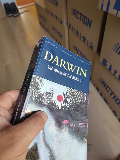 Image of moving boxes in the background and me holding up a book on the foreground. The boon is "the voyage of the Beagle" by Charles Darwin.
