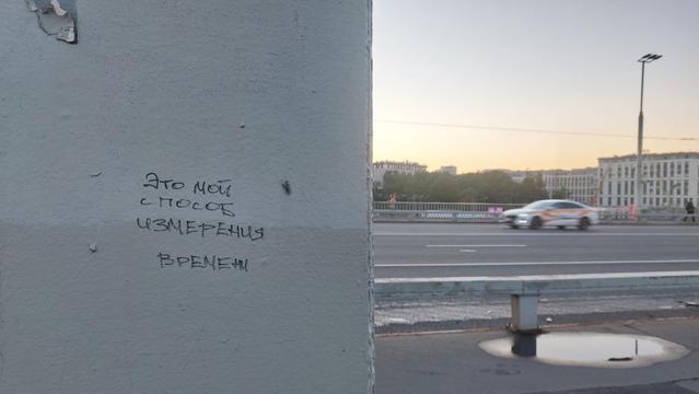 Фото надписи «Этой мой способ измерения времени» на опоре моста, которая занимает левую половину фото. На правой половине фото проезжая часть и краны на стройке ЖК рядом с офисным зданием.