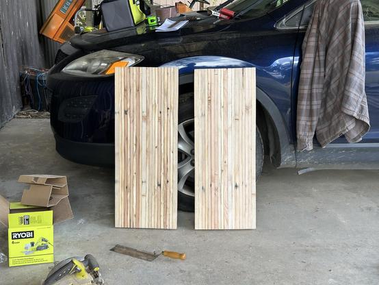 Two bench tops leaning against a car. They’re butcher-block style, lots of long slats. The car’s hood has a bunch of tools on it, and a jacket is hanging from the side view mirror. There is a hand saw and a circular saw on the floor, along with a box for another tool.
