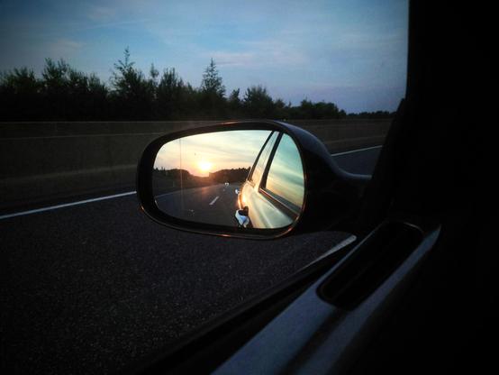 Zu sehen ein Blick in den Seitenspiegel eines Autos am Morgen vom Fahrersitz aus. Man sieht dunkel ein Stück der tür im Fenster dunkel die Straße und bäume am Rand und blauen Himmel darüber. Im Spiegel sieht man golden die Sonne hinter dem Auto der Himmel ist hell. Die Sonne spiegelt sich auch in der Fahrertür.