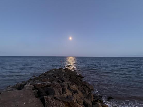 Photo of a not quite full moon hanging over the sea in a mid blue summer night sky. Its silver-gold reflection beams across the rippled surface towards a rocky groyne that juts out from the shore in the foreground. Waves break gently and foam white on the right. The sea is an inky blue.