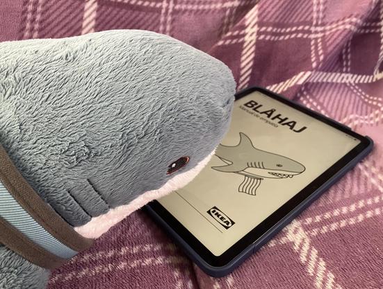 A Blåhaj cuddly shark looking at a tablet displaying an Ikea Blåhaj “Manual de empatía” instruction leaflet.