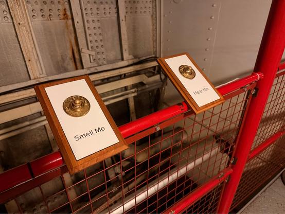 there are two brass knobs on plaques mounted to a red metal railing. one is labelled “Smell Me” and the other is labelled “Hear Me”