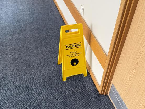 a freestanding yellow warning sign on a hotel floor says “Caution: Robotic vacuum cleaners in operation”