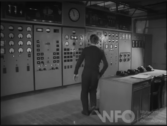 Frame from a black and white movie from the 60s, showing the control room of a large factory, with a full wall of dials and buttons.