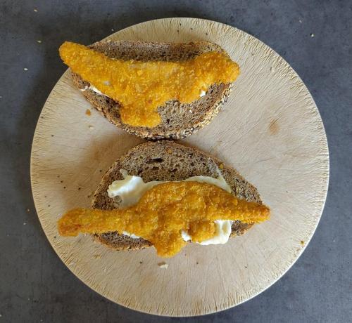 A photo showing a bread board with a sandwich being prepared. On the bread are two dinosaur shaped nuggets, with some mayonnaise.