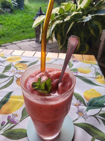 Eine Nahaufnahme zeigt ein Glas mit einem dicken, rosa gefärbten Getränk auf einer gemusterten Tischdecke. Im Getränk befinden sich ein orangefarbener Strohhalm, ein Metalllöffel und ein kleiner Zweig grüner Minze. Das Glas steht auf einem hellblauen Untersetzer auf einer weißen Tischdecke mit gelben Zitronen, grünen Blättern und kleinen Blüten. Der Hintergrund zeigt einen Garten mit grünem Rasen, Steinplatten und großblätt großblättrigen Pflanzen. Ein dunkles, laternenartiges Objekt ist hinter dem Glas sichtbar.