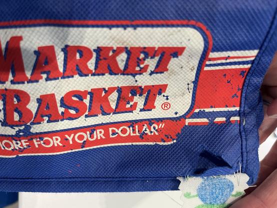 A blue reusable grocery bag with a very scraped up Market Basket logo on it. In the bottom corner of the bag is a triangular rip. A patch has been half-sewn on because I always forget to take photos before I start working 🤦‍♀️
