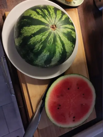 An overhead view of half of a green striped watermelon on a white plate, sitting next to a sliced round piece of watermelon with bright red flesh and black seeds on a wooden cutting board with a kitchen knife.