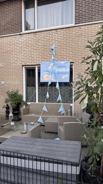 On the front of a house, a light blue poster is hung to announce the birth of a baby boy. Blue bunting hangs drooping from the facade. In the garden some cheap grey garden furniture, a table with multiple ashtrays and a packet of Marlboro. The poster shows an angelic sleeping baby and the text, in Dutch: “Hurray! Diablo is born!”